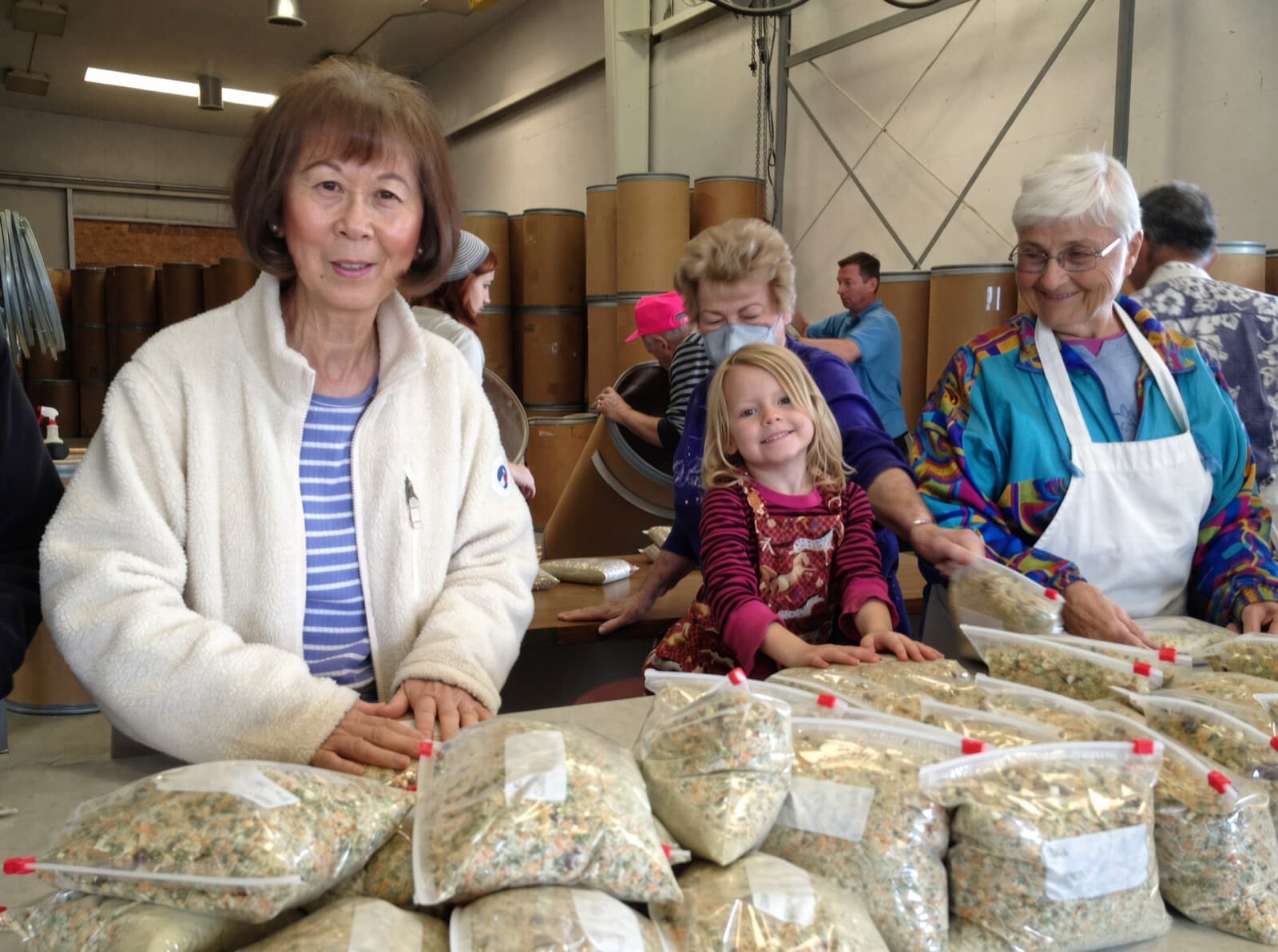 Volunteers of all ages, from a young child to older adults, smiling together as they pack bags of food at a community serving event